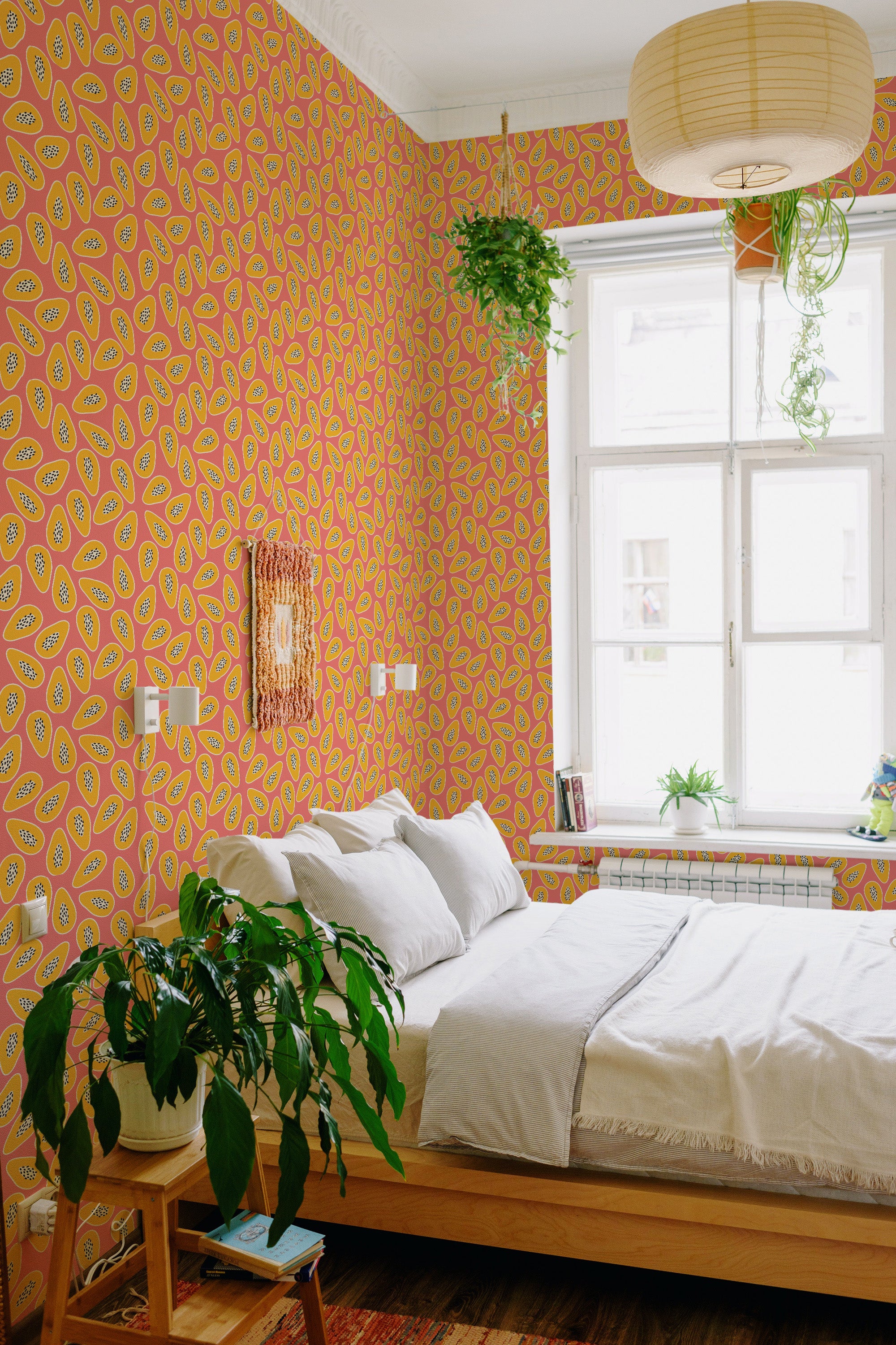 A room with vibrant yellow lemons and green leaves on a pink background covers the walls, creating a lively and playful vibe. The pattern is repeated across the entire wall, with white wooden window frames allowing natural light into the space. The room is cozy with a bed, woven wall art, and various potted plants adding to the warmth. The wallpaper design brings a fresh, nature-inspired element to the traditional wallpaper.