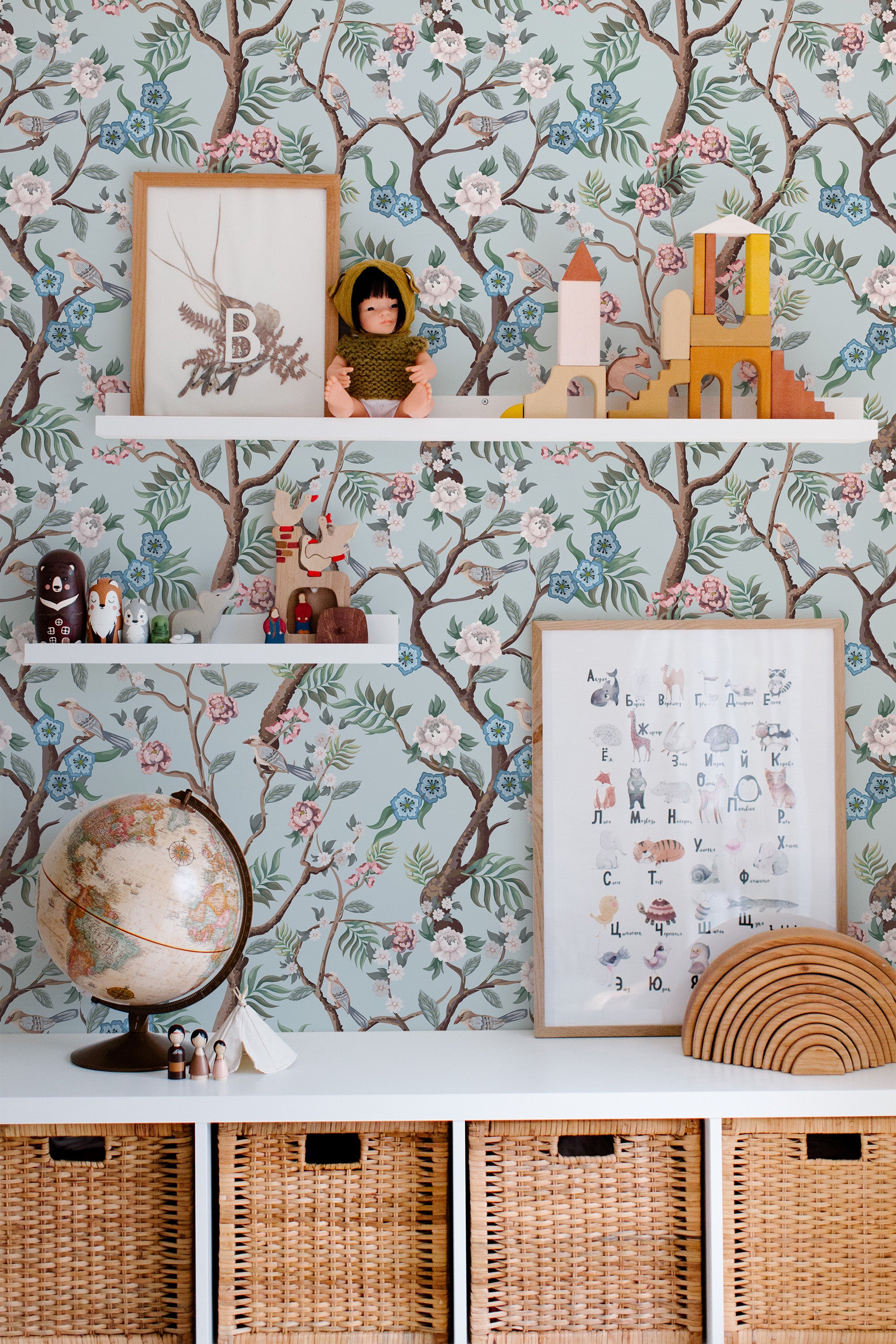 Self-adhesive wallpaper with a tree design, featuring intricate branches, delicate pink flowers, and green leaves set against a light blue background. The natural elements create a serene, nature-inspired look in the room, complementing the decor on the shelves and the wicker baskets below.