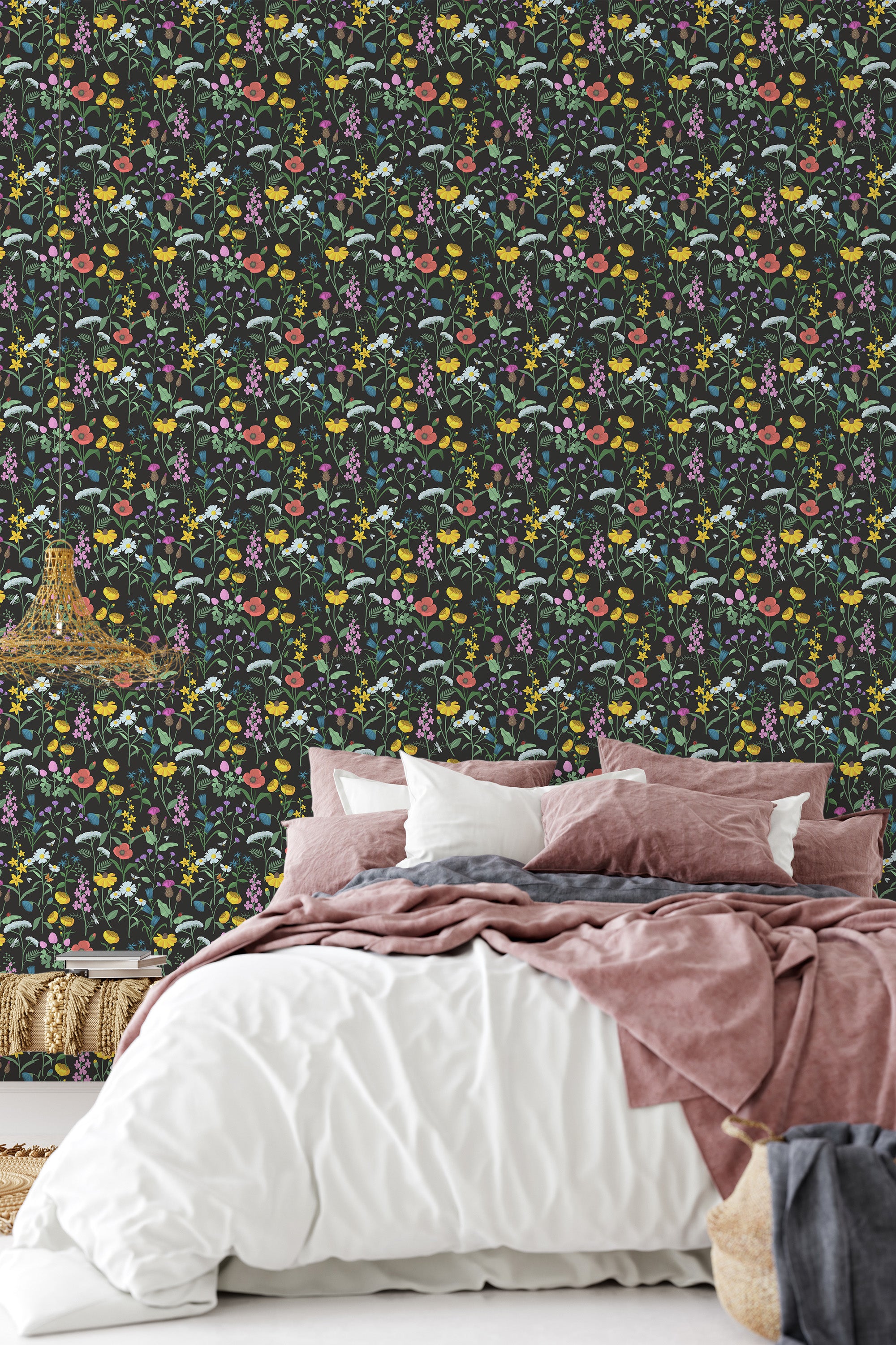 A bedroom wall covered in a dark Meadow wallpaper design with a repeating pattern of yellow lemons, green leaves, pink and white flowers, evoking a vintage garden feel. Below, a bed is adorned with soft, neutral-colored bedding. The wall has a striking contrast with the bedding, creating a cozy and traditional wallpaper setting.
