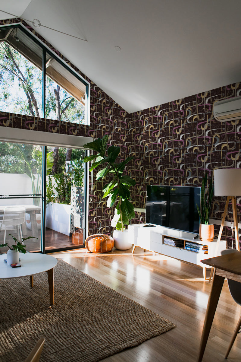 A room with walls covered in a colorful brush stroke patterned wallpaper, blending hues of purple, yellow, and beige. The vibrant design contrasts with the modern decor, complementing the natural light from large windows and the greenery inside the space. Traditional wallpaper.