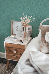 Self-adhesive modern wallpaper in a geometric pattern of intersecting lines and diamond shapes on a teal background above a wooden nightstand with books, a picture frame, and a vase of dried flowers.