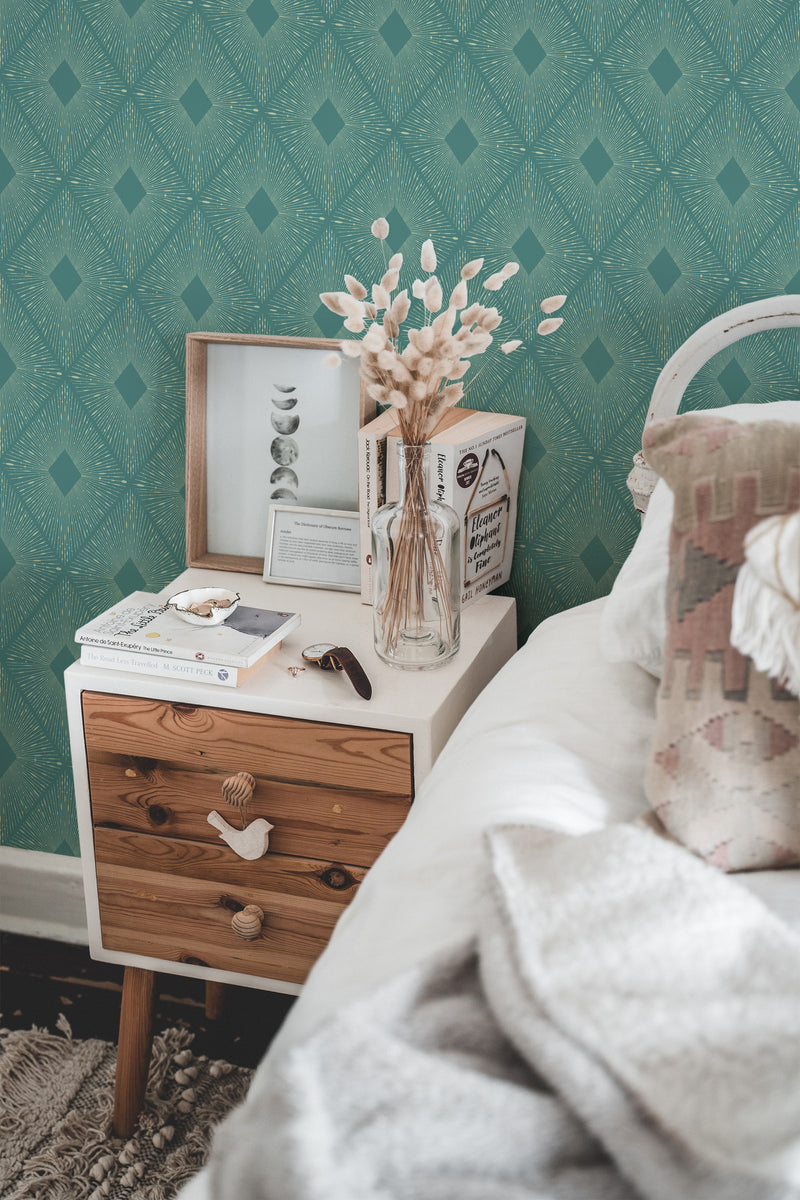 Self-adhesive modern wallpaper in a geometric pattern of intersecting lines and diamond shapes on a teal background above a wooden nightstand with books, a picture frame, and a vase of dried flowers.