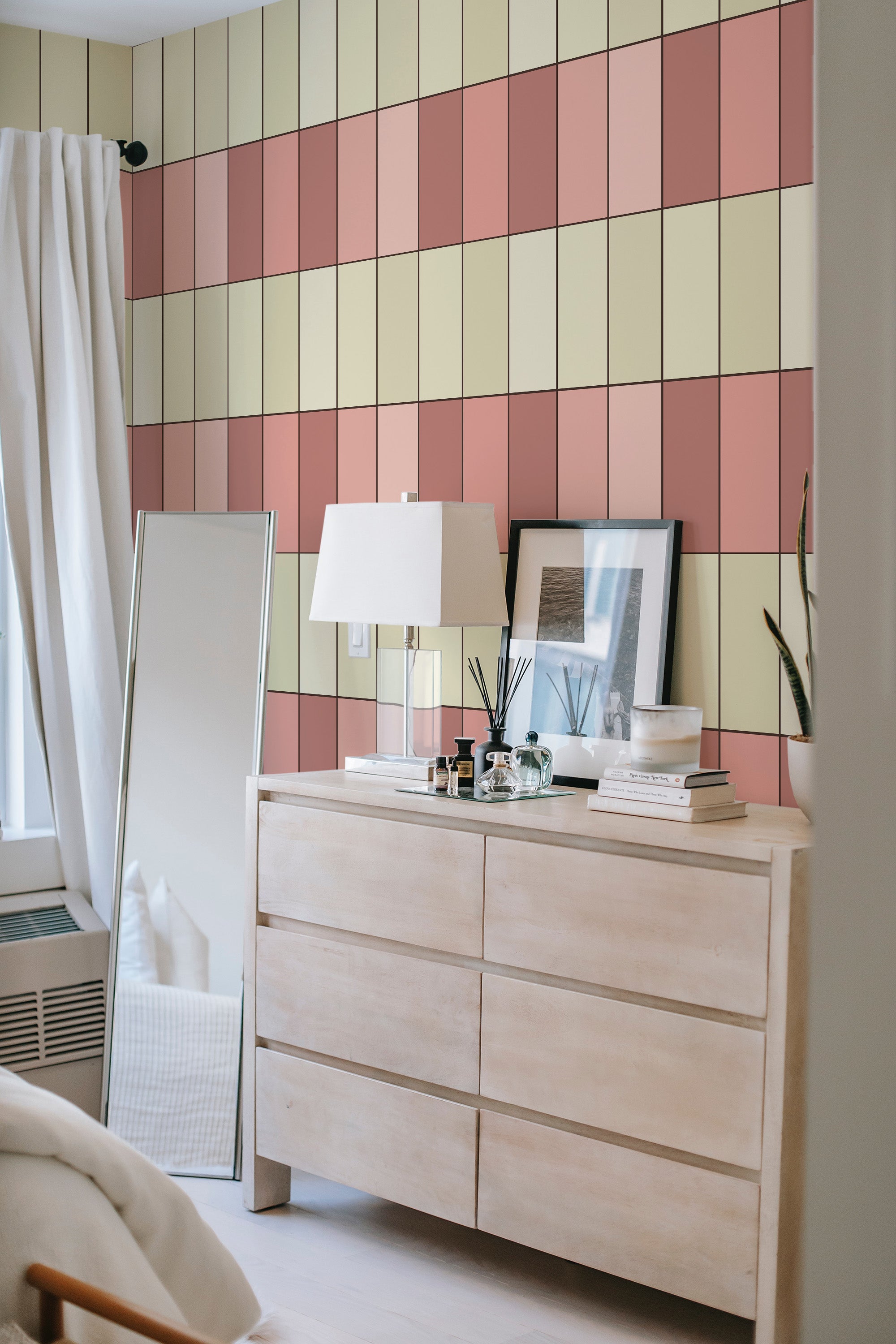 An interior room with a rustic wooden dresser and large mirror next to a wall covered in wallpaper with vertical skinny tiles in shades of pink, peach, and cream, creating a geometric pattern for a modern take on traditional wallpaper.