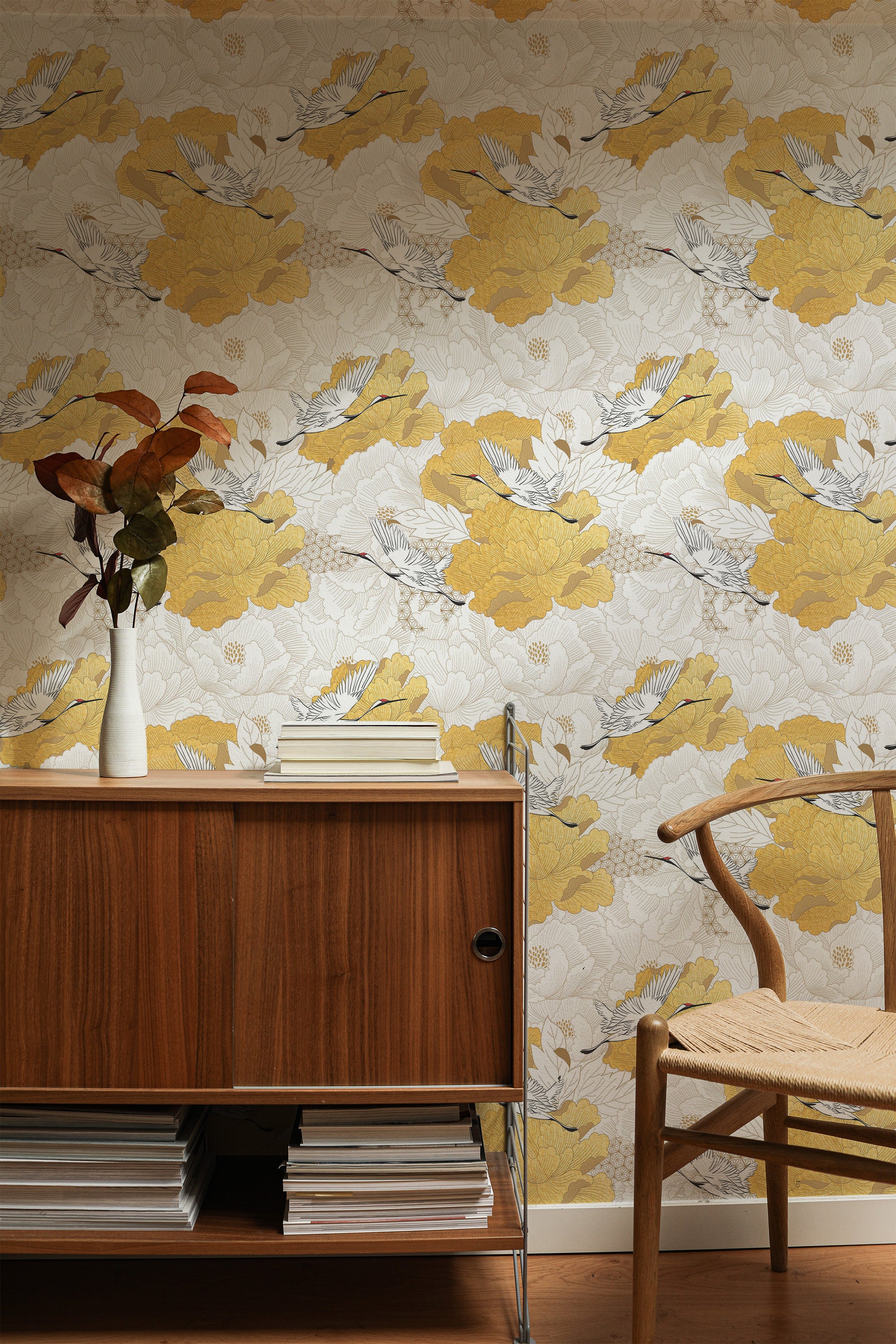 Self-adhesive wallpaper with a Chinoiserie bird design in a palette of yellow, gray, and white, set against a wooden sideboard and chair, complemented by a vase with autumn-colored leaves.