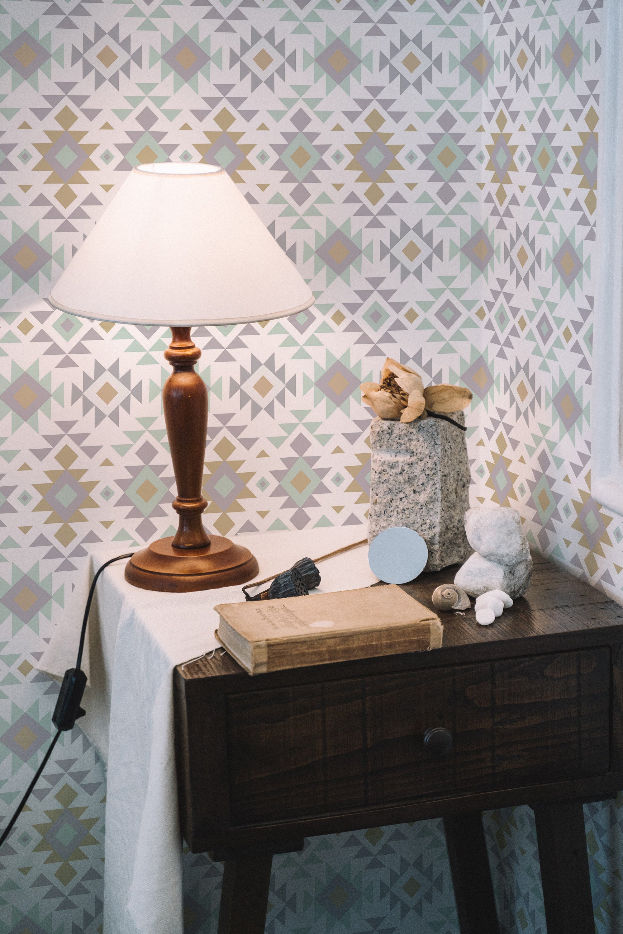 Self-adhesive pastel Aztec wallpaper with geometric patterns in soft shades of gray, beige, and muted mint green, enhancing a cozy room with a wooden side table, a lamp, and decorative items.