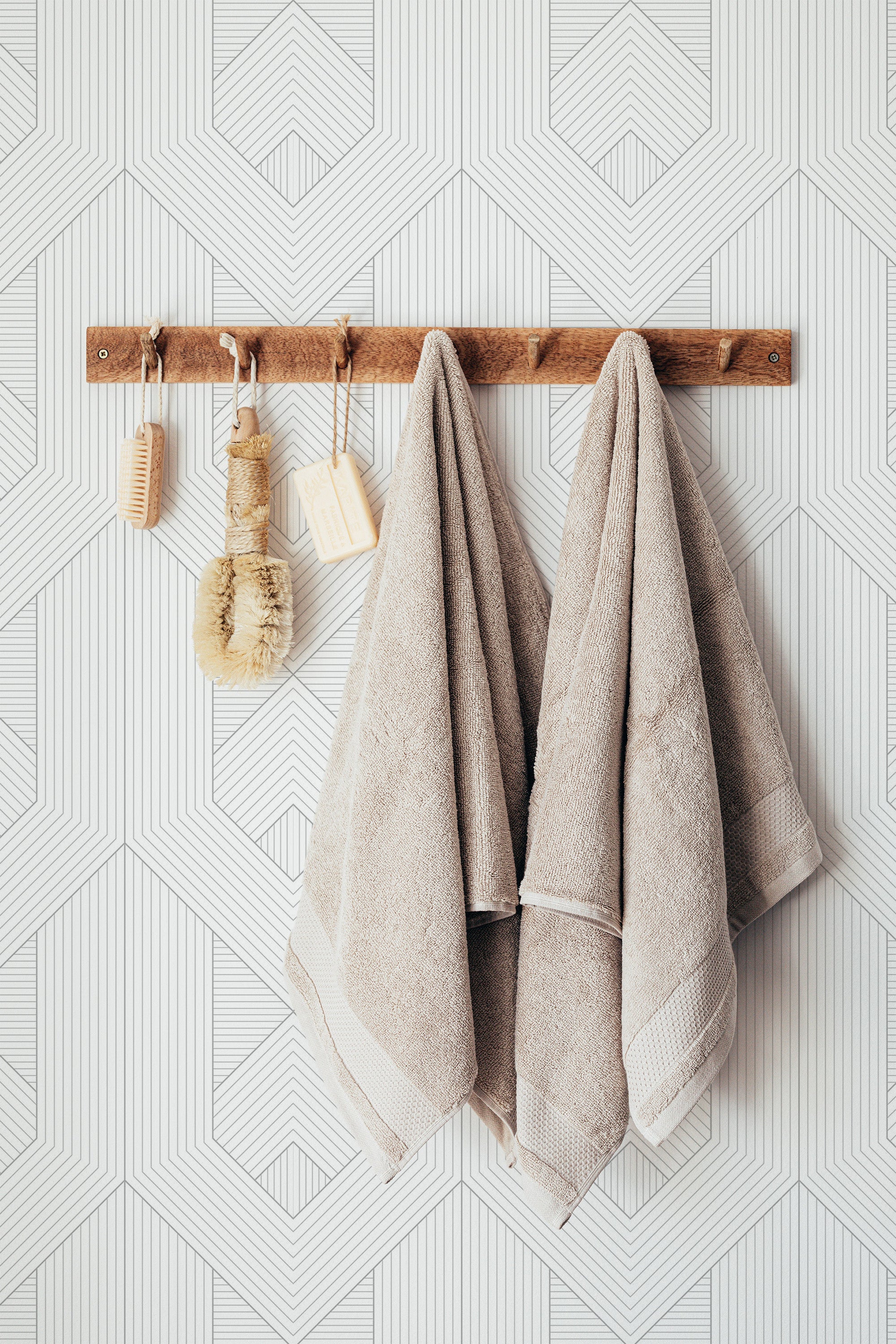 Self-adhesive minimalist wallpaper with a geometric line art pattern in light, neutral tones, rectangular shapes intersect throughout, creating a subtle, modern visual interest on the wall behind a wooden towel rack with hanging beige towels and rustic bath accessories.
