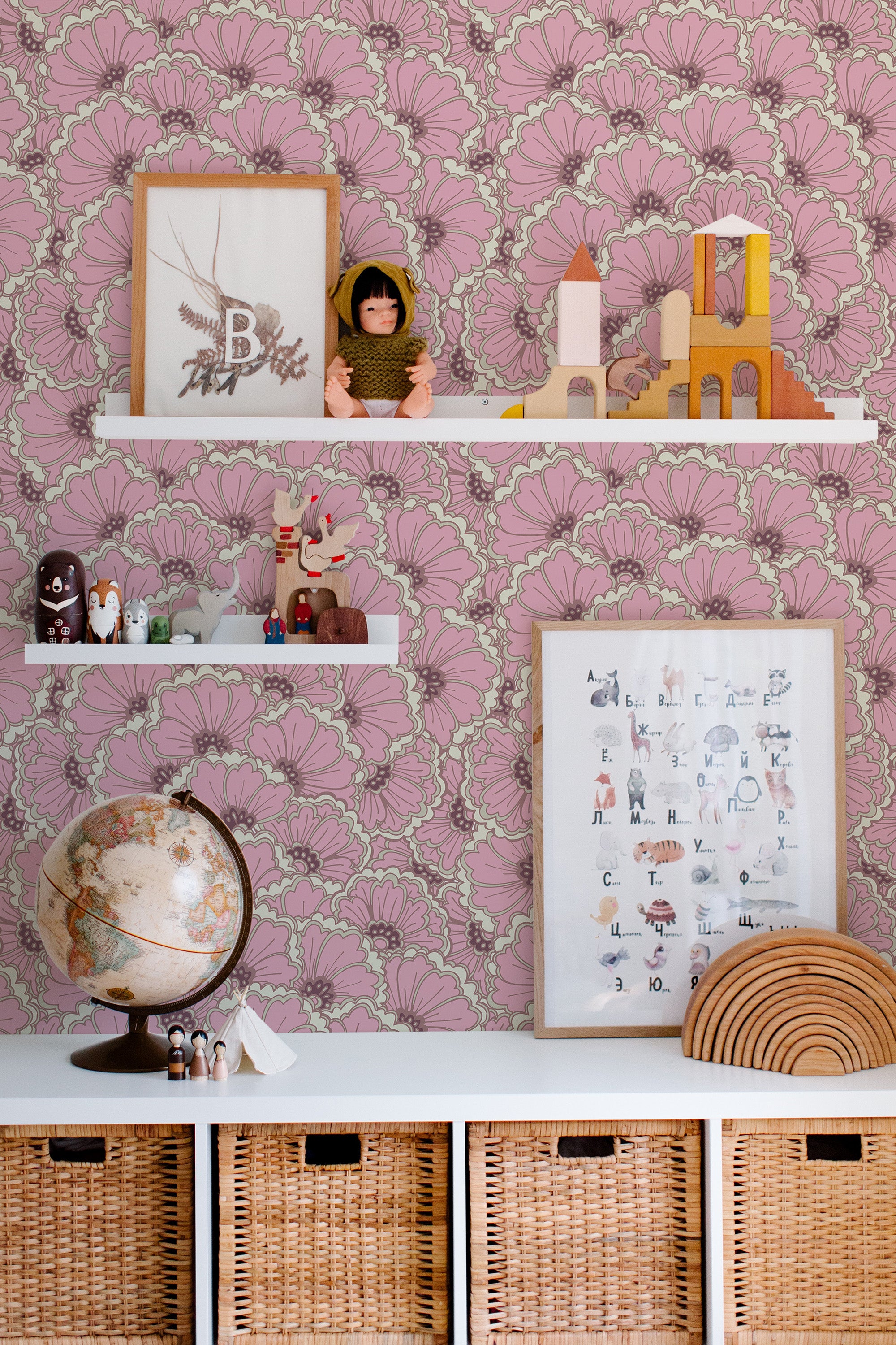 Self-adhesive wallpaper with a bold eclectic print in shades of pink featuring large intricate floral patterns behind white shelves displaying toys, framed art, and a globe.