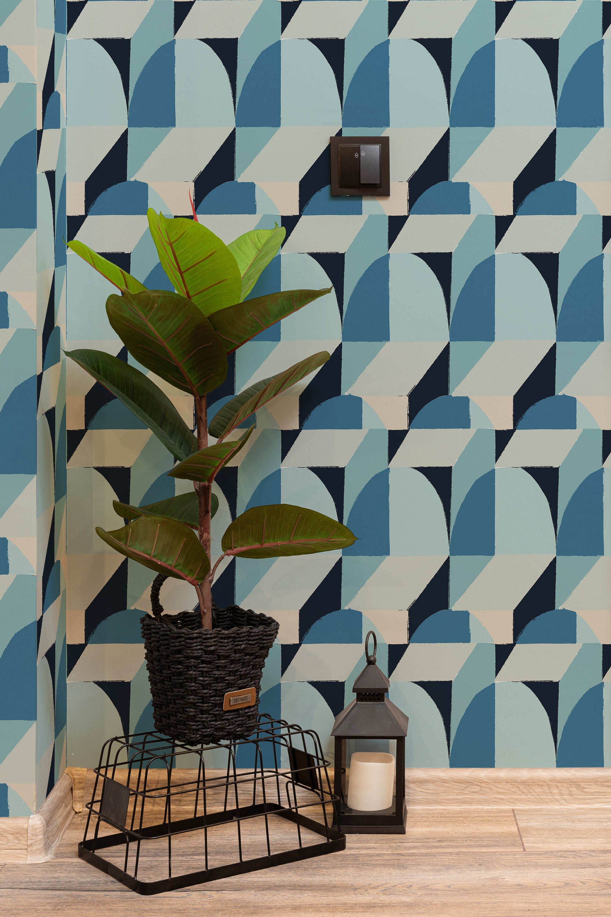 Self-adhesive wallpaper with a Blue Abstract Cubes design, featuring geometric blue and beige patterns with a 3D effect. The wallpaper covers a wall in a modern room with wooden floors, complemented by a potted plant, black lantern, and light switch as accents.