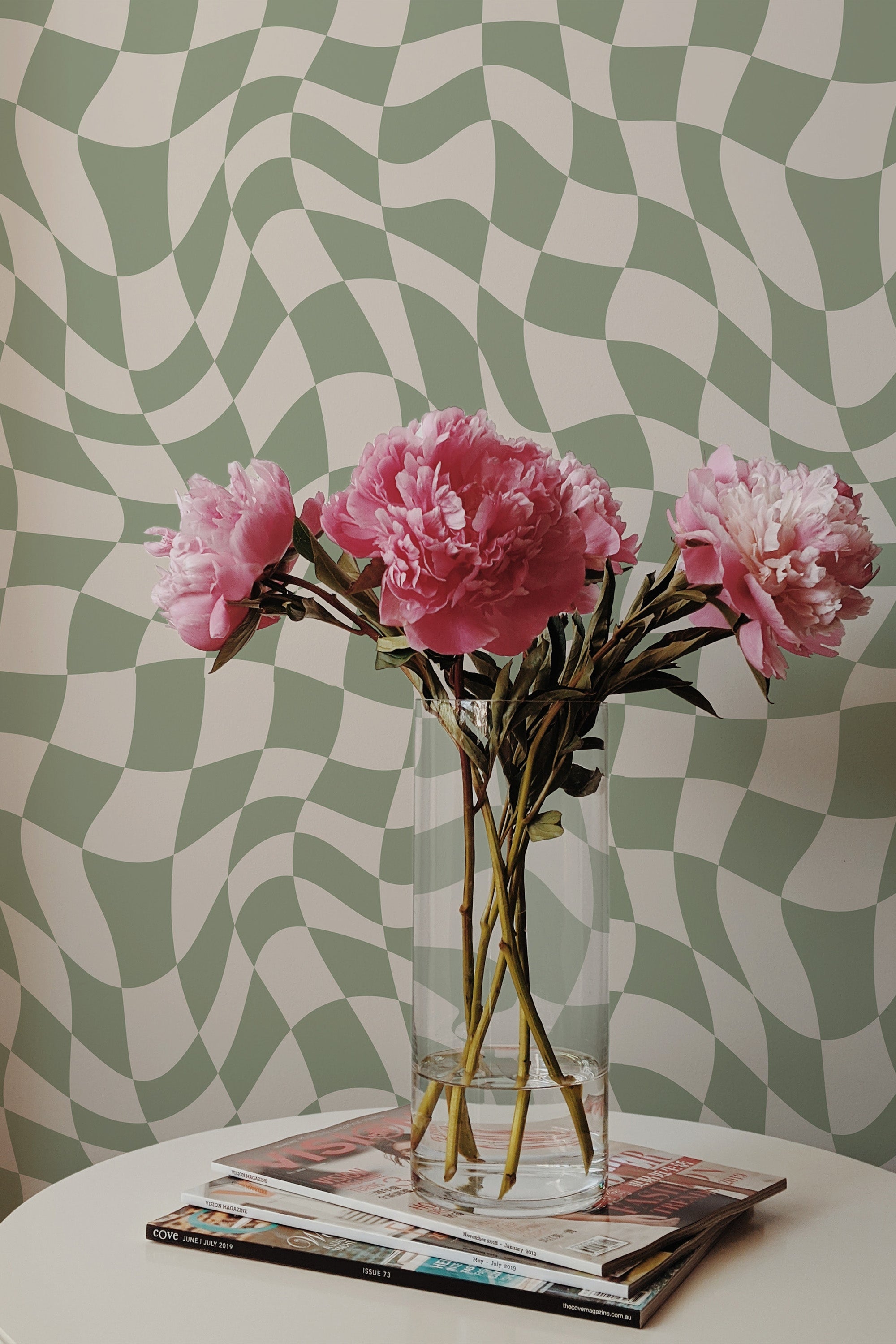 Self-adhesive wallpaper with a funky checker pattern in shades of green and off-white, creating a wavy optical illusion effect behind a vase of pink peonies on a table.