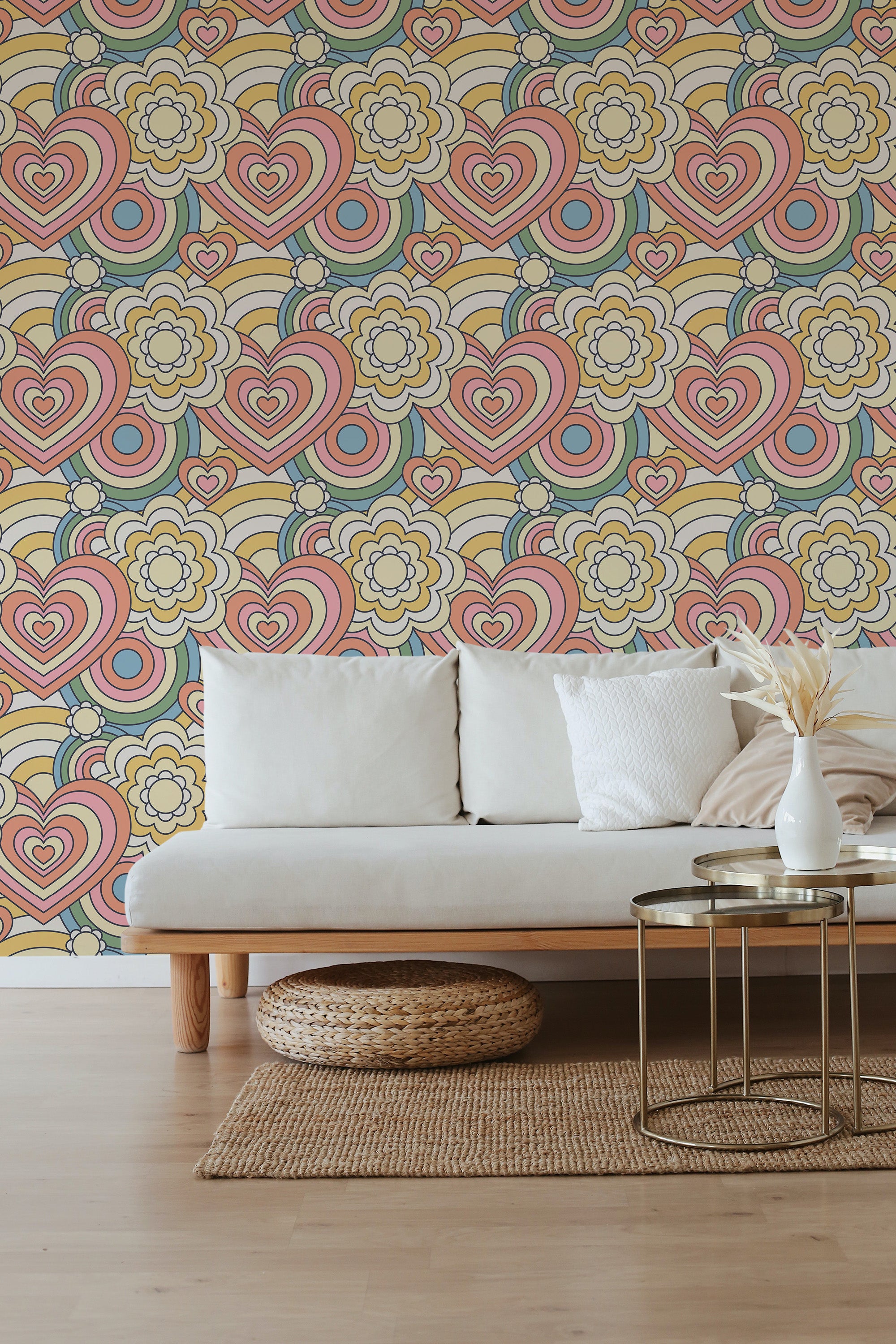 A room with a beige sofa, rattan pouf, and glass coffee table. The wall has a pastel retro wallpaper with hearts, flowers, and geometric shapes in shades of pink, yellow, and blue. Traditional wallpaper.