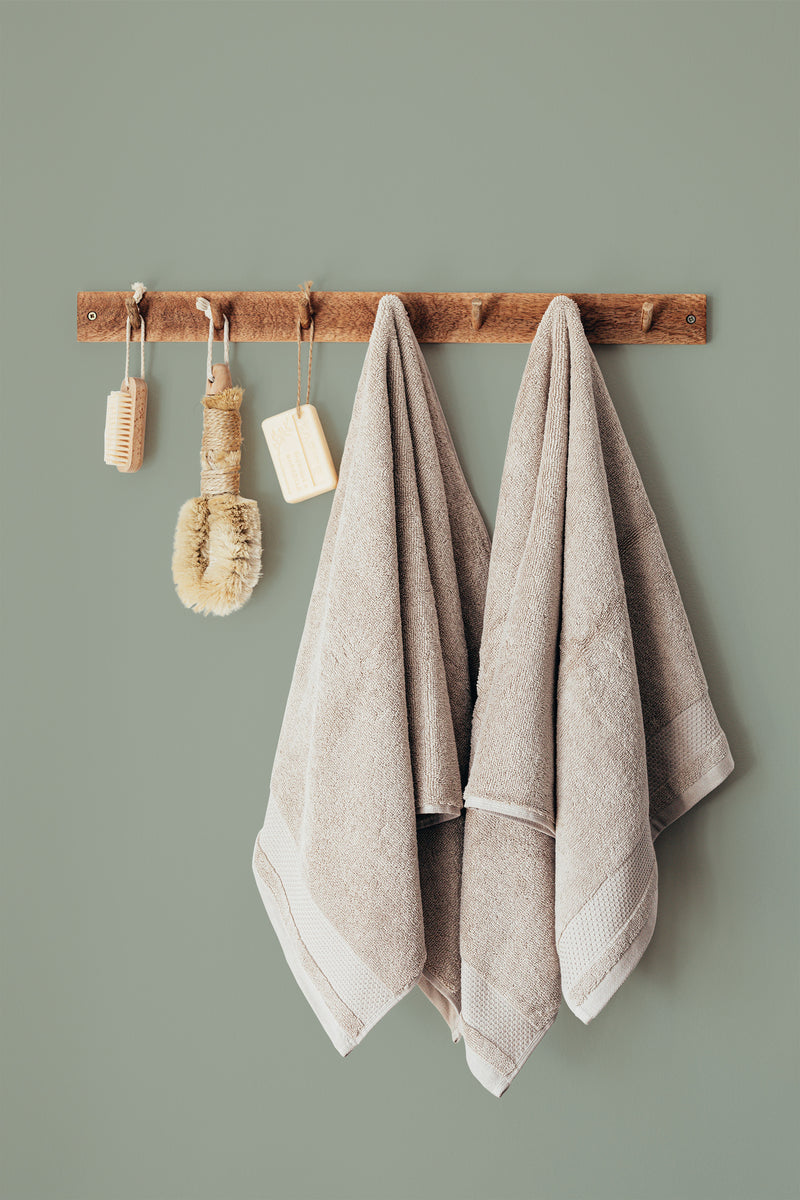 Self-adhesive wallpaper in a muted sage green solid color installed on a wall, creating a calm and soothing backdrop. A wooden rack with beige towels and bathroom accessories hangs against the wallpaper.