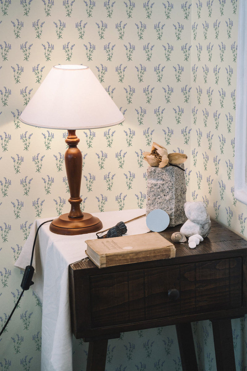 Self-adhesive wallpaper with a delicate lavender pattern on a light background, covering the walls of a cozy room with a rustic wooden table, a beige lampshade, books, a plush toy, and other decorative items.