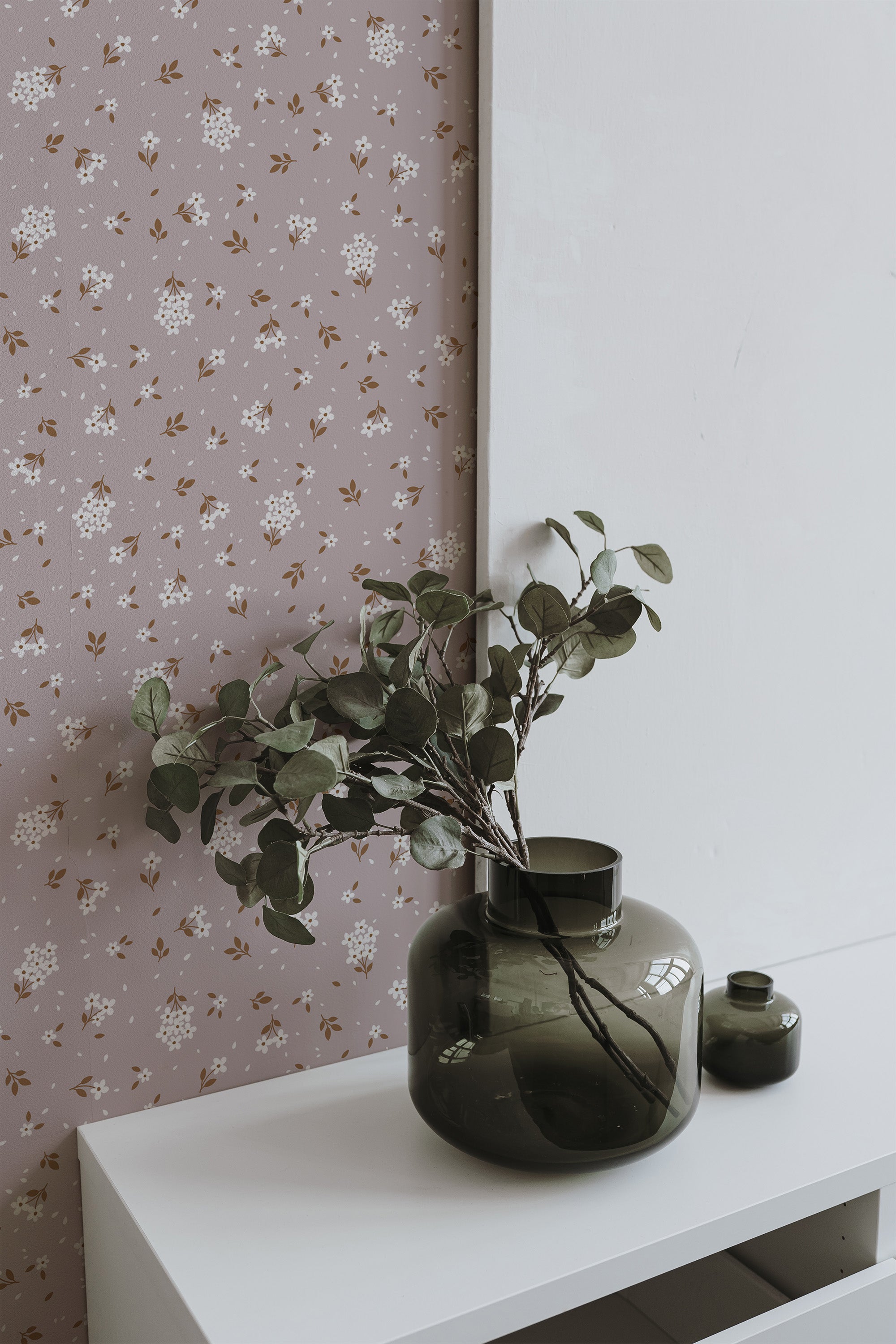 Self-adhesive wallpaper with a pattern of pinkish delicate flowers and subtle green leaves on a beige background, partially covered by a white canvas and accompanied by green vases with foliage.