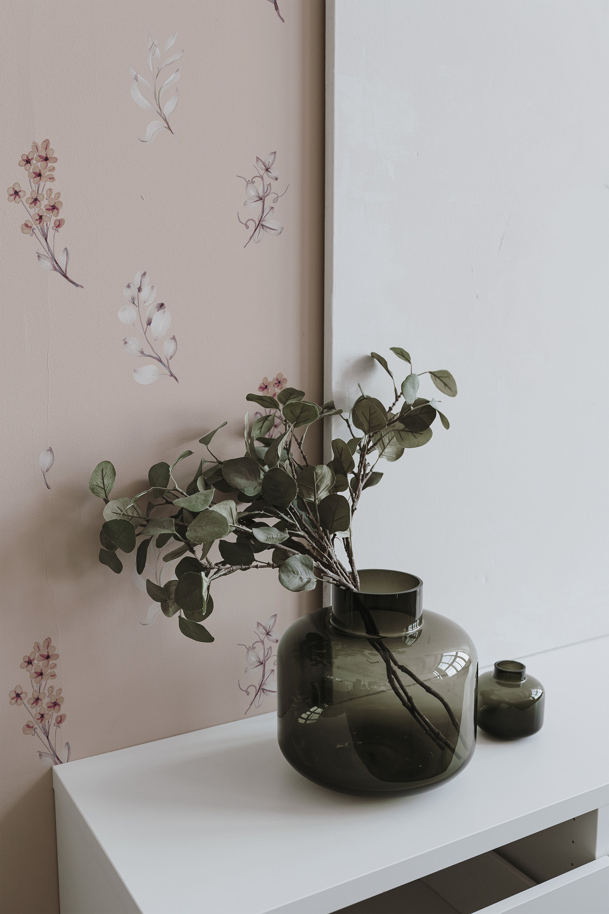 Self-adhesive wallpaper with a flying flowers design, incorporating subtle florals and leaves in muted tones on a light background, in a modern styled room with a vase on a white shelf.