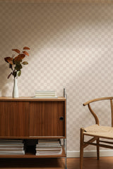 Self-adhesive Neutral check pattern wallpaper in light beige hues applied on a wall behind a wooden sideboard and a chair with a plant and books on top.