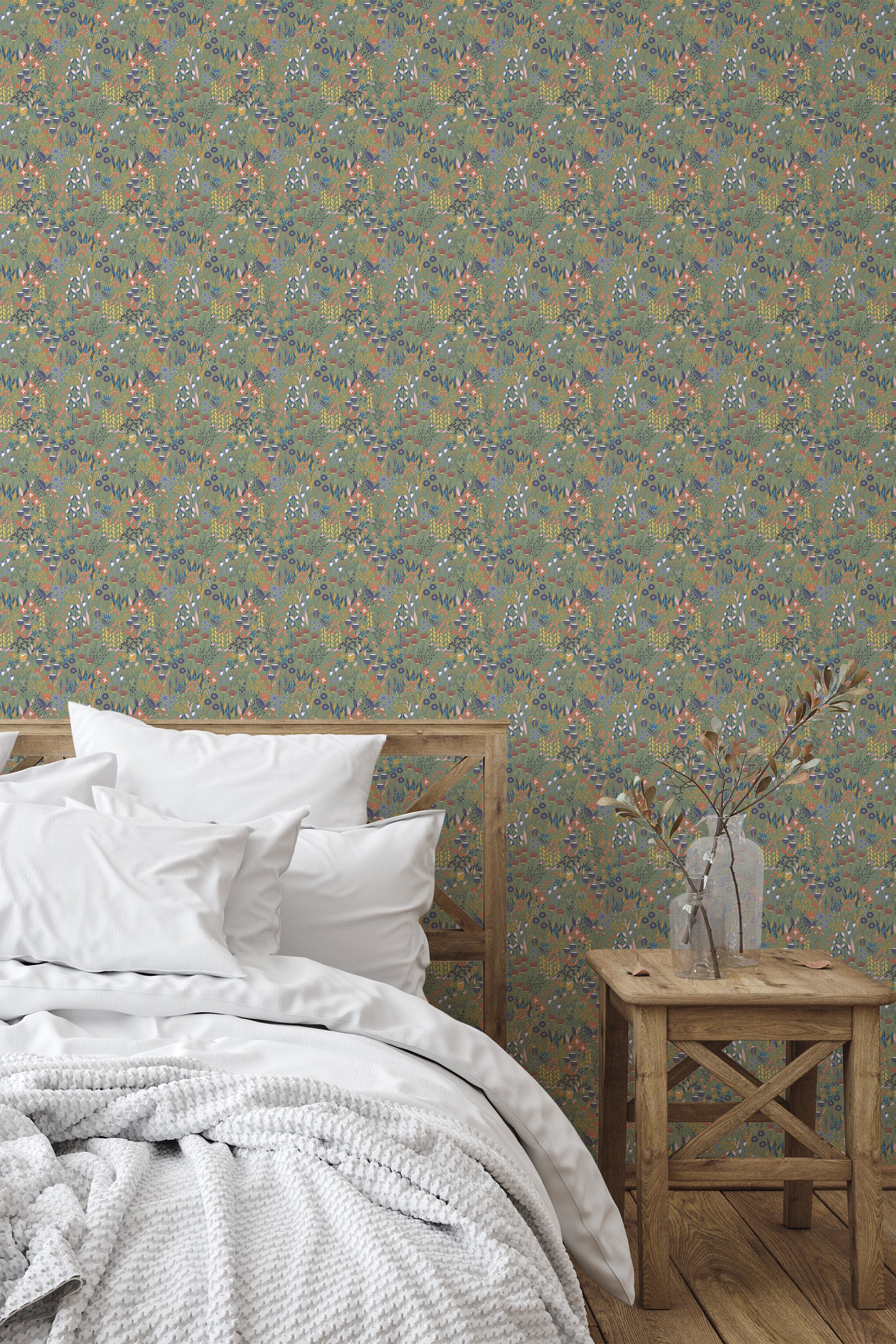 Self-adhesive Sage Meadow wallpaper with a pattern of various plants and flowers in soft green, blue, and pink hues, covering a bedroom wall behind a wooden bed and side table.