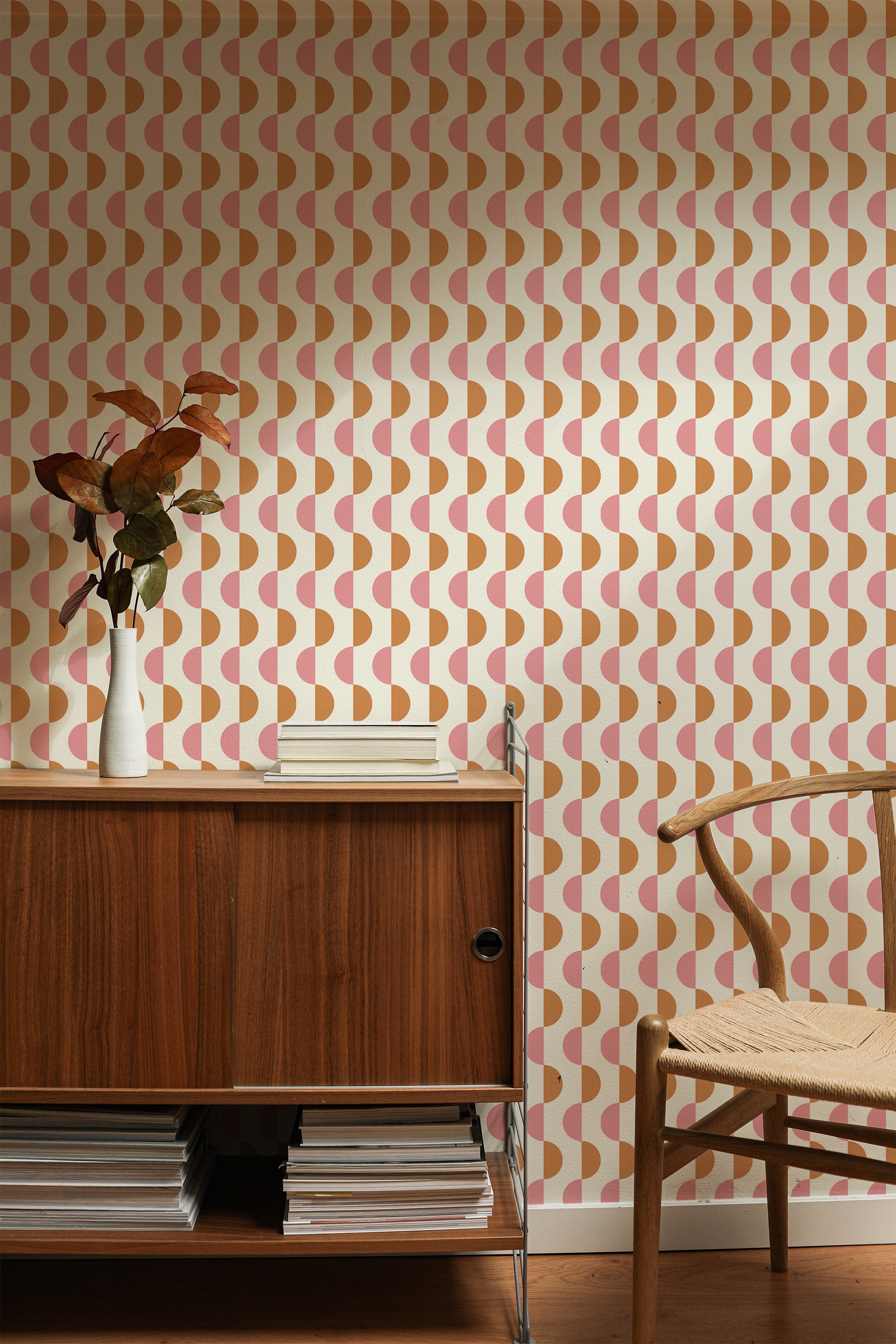 Self-adhesive wallpaper with a mid-century half circles design in peach and cream colors on a wall behind a wooden credenza and chair, complemented by a vase with dried plants.