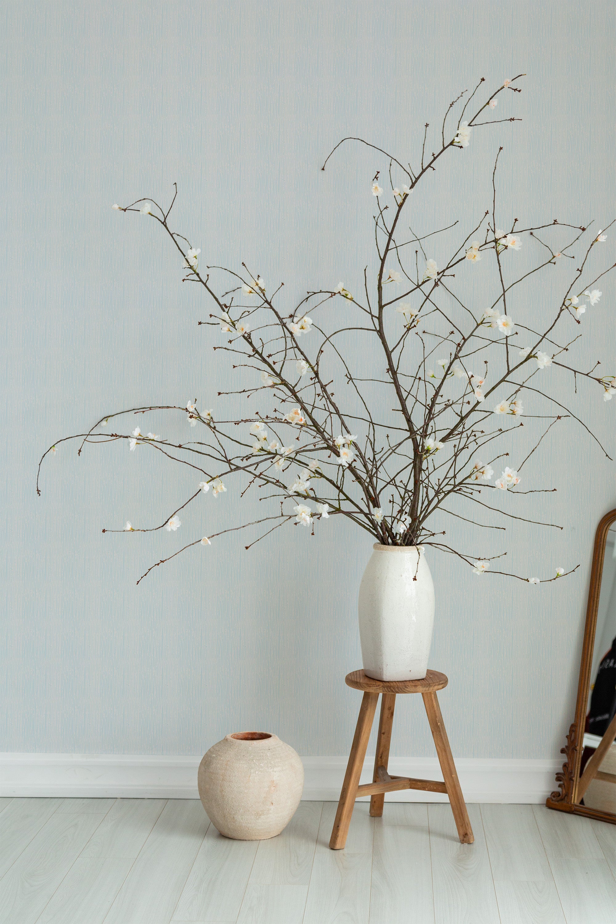 Self-adhesive wallpaper with a subtle Rainy Day design on a light-colored wall, accompanied by a vase of blossoming branches on a wooden stool.