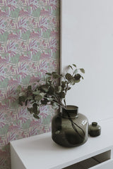 Self-adhesive wallpaper with a leaf and flower pattern in hues of pink and green on a wall, accompanied by a white shelf and decorative green vases with foliage.