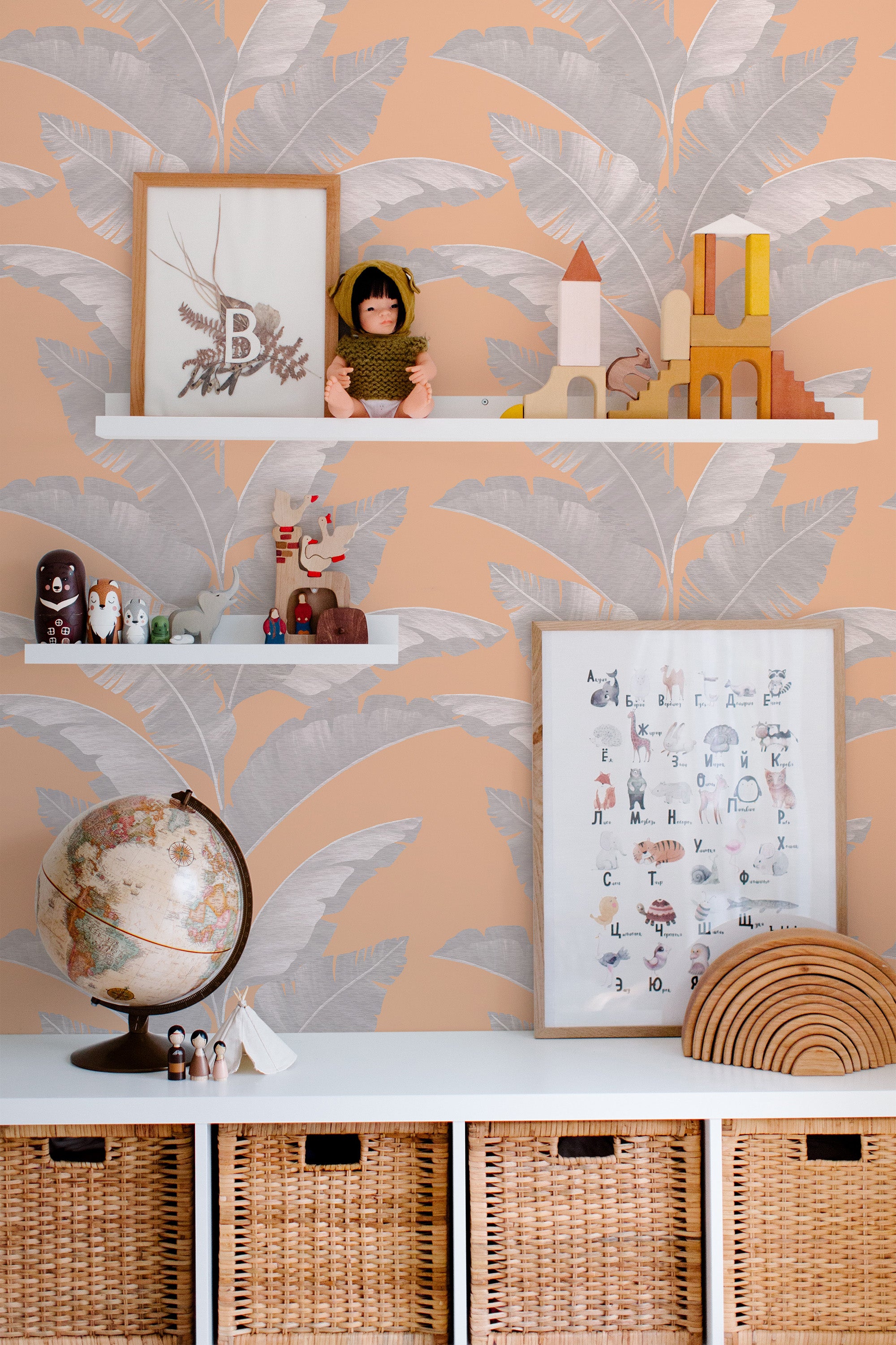 Self-adhesive Peachy Tropical wallpaper with large grey and white leaf patterns on a peach background in a room with children's toys and educational posters.