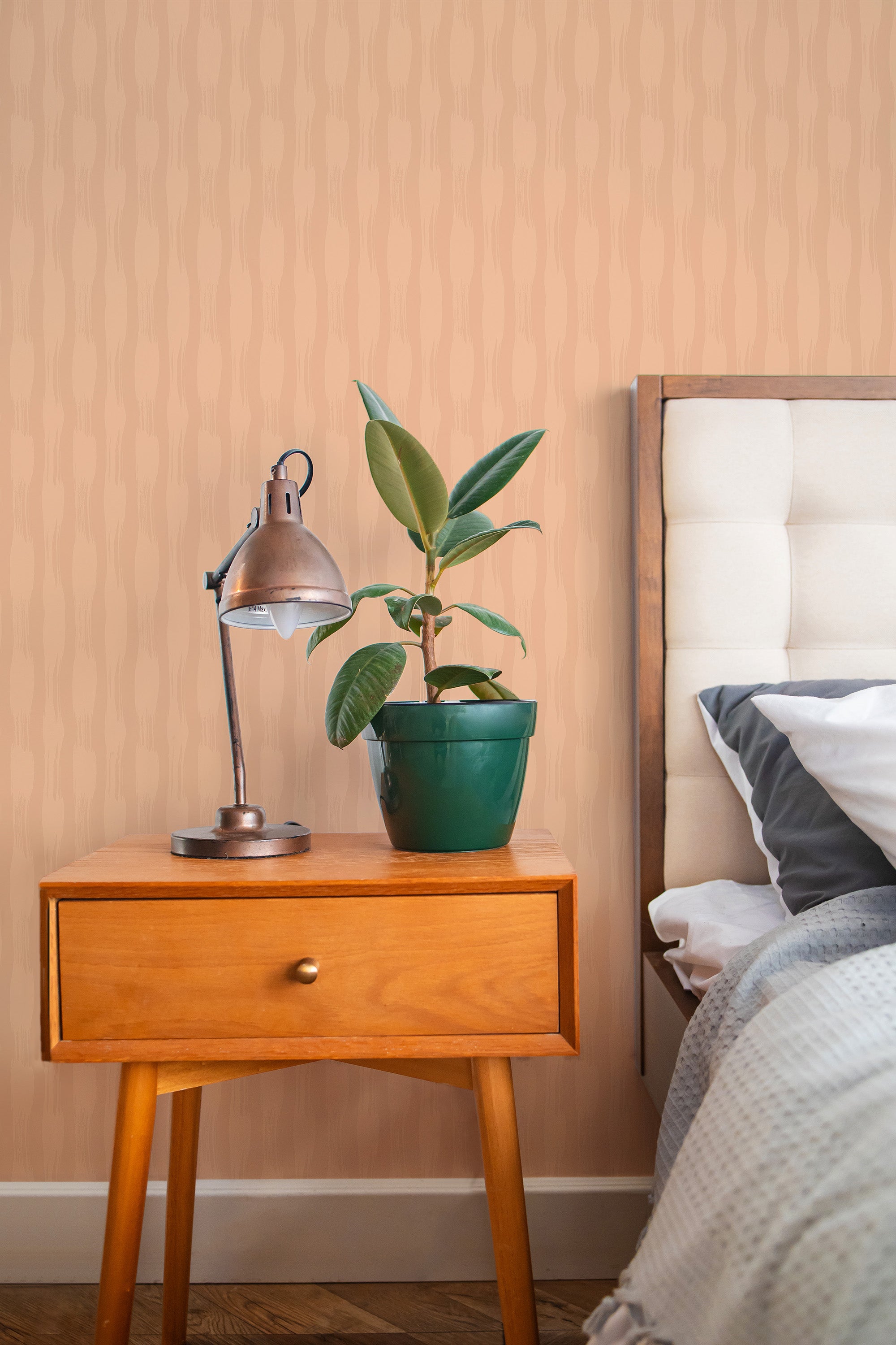 Self-adhesive Peach Brush Strokes wallpaper with vertical pink and cream pattern on a wall in a bedroom with a wooden bedside table, lamp, and potted plant.