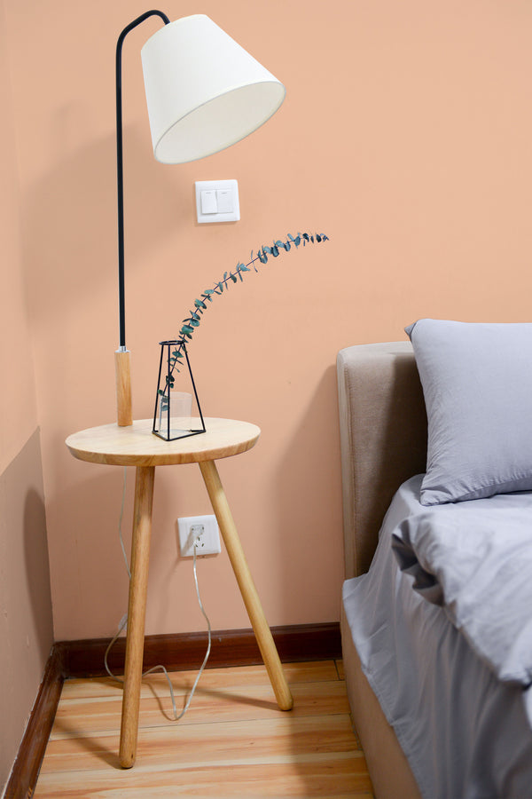 Self-adhesive solid color peach wallpaper on a room wall with a bedside table, lamp, and part of a bed visible.