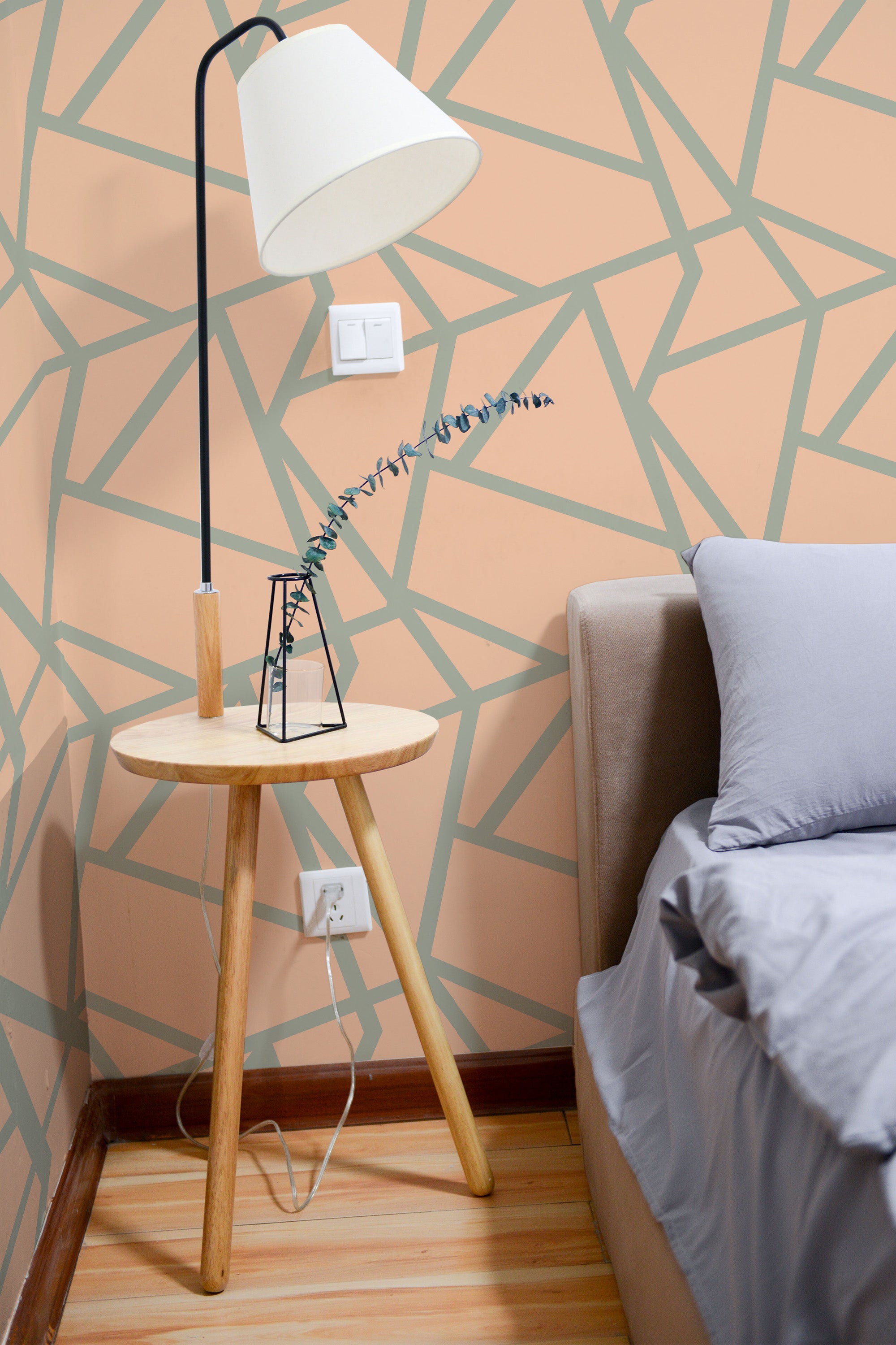 Self-adhesive wallpaper with peach and green geometric lines on an off-white background in a cozy room with a floor lamp, side table, and part of a bed visible.