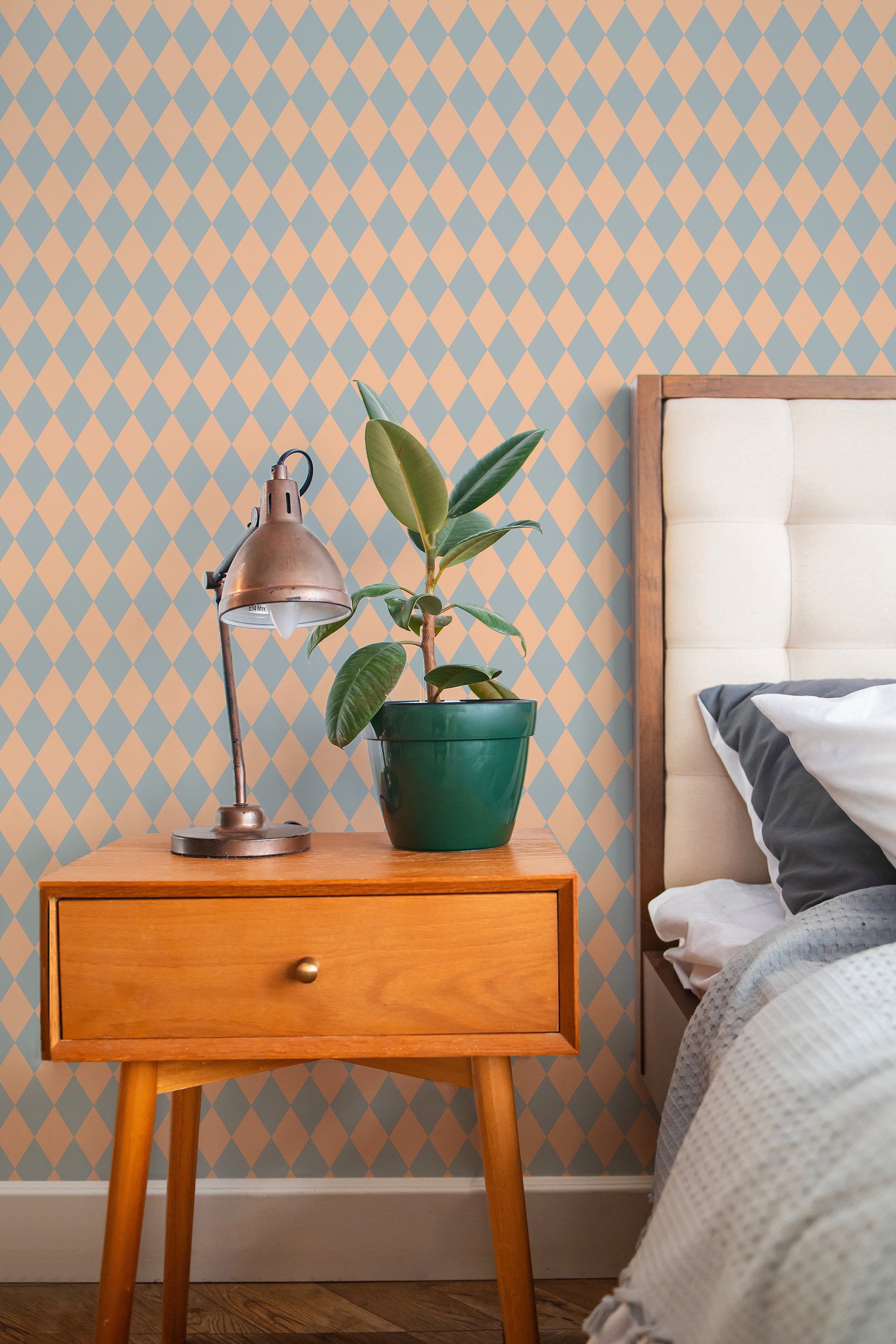 Self-adhesive Peach and Sage Harlequin wallpaper with diamond pattern on a room wall beside a wooden bedside table with a lamp and plant.