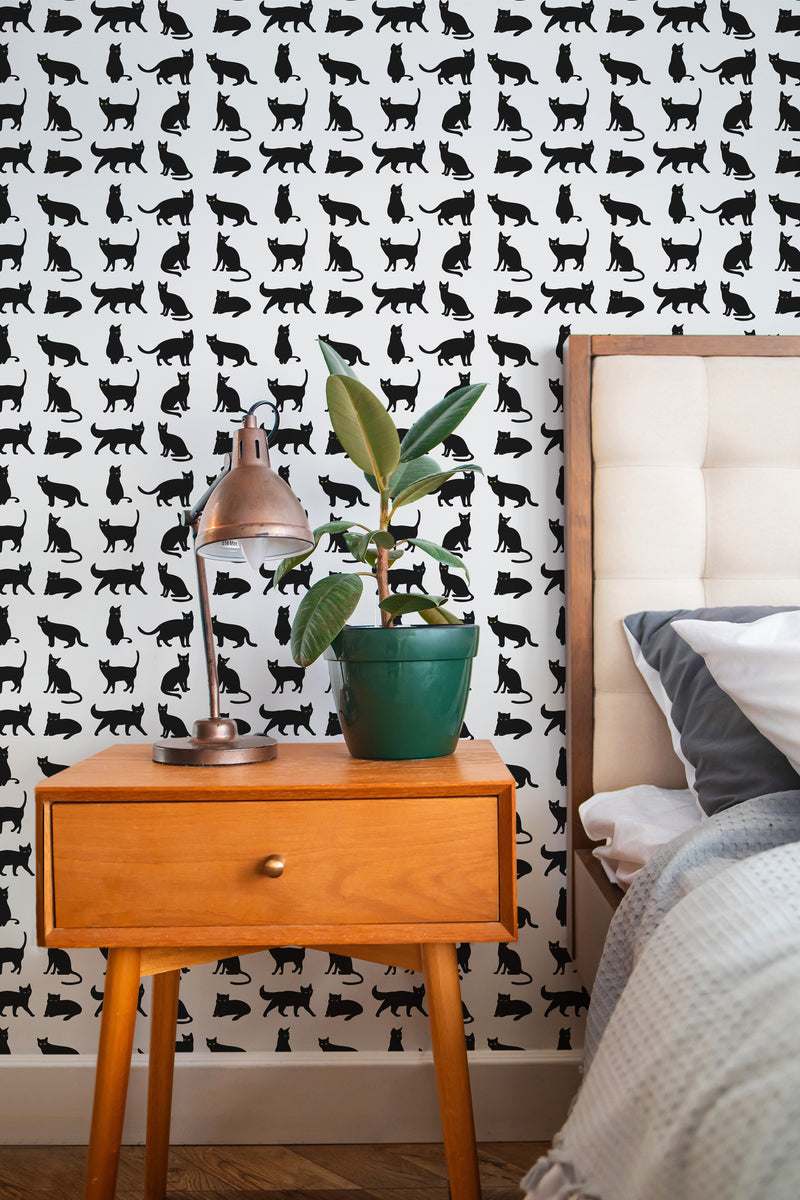 Self-adhesive wallpaper with a repeating pattern of black and white stylized cats in various poses on a wall behind a wooden bedside table with a plant and lamp.