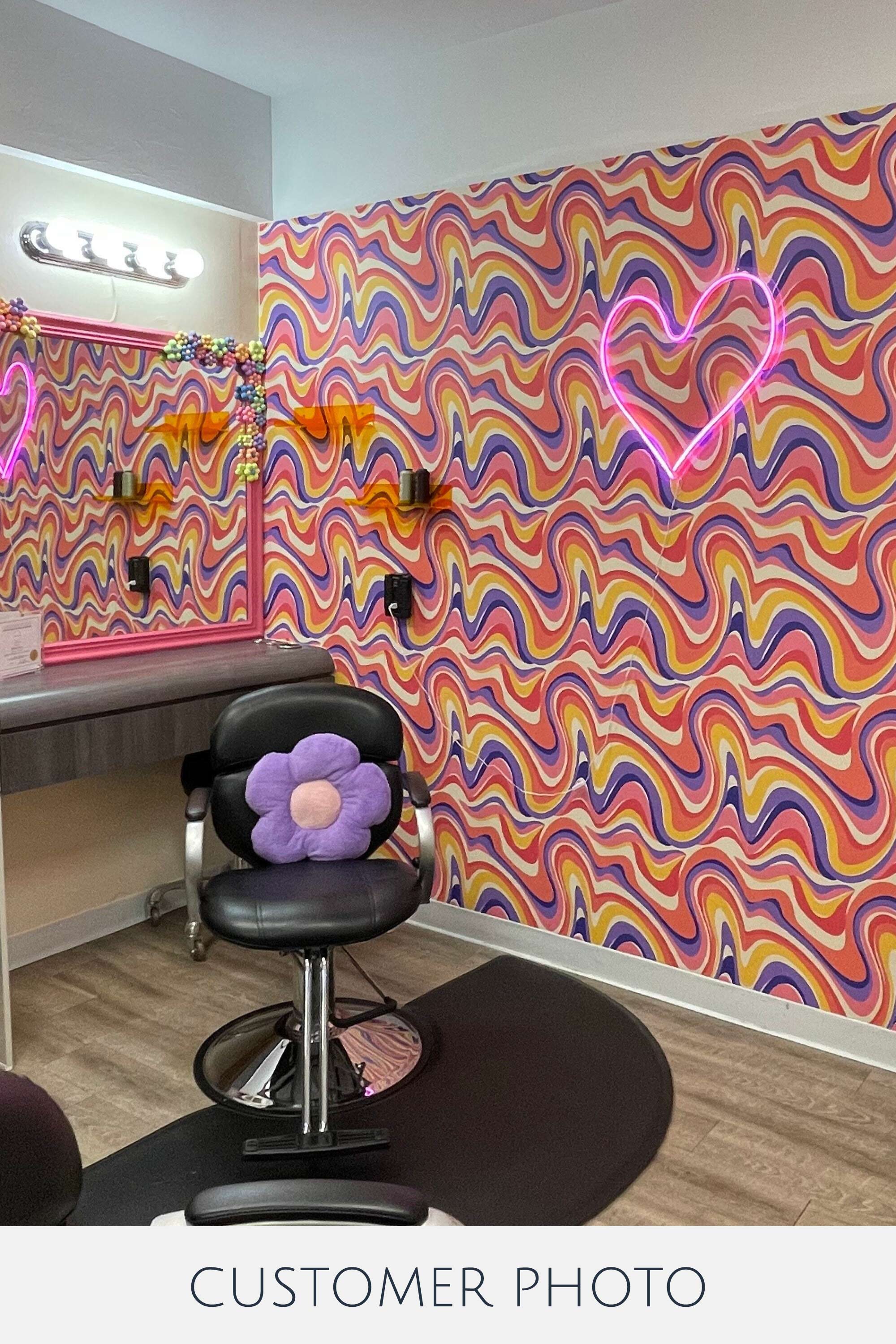 Psychedelic wallpaper with vibrant, wavy patterns in pink, orange, purple, and yellow, evoking a sense of movement and energy. A neon pink heart light hangs on the wall, adding a modern twist to the colorful, swirling design. The room includes a black salon chair with a purple flower cushion, wooden flooring, and a mirror with illuminated bulbs, creating a stylish contrast to the lively wall pattern. This is traditional wallpaper.